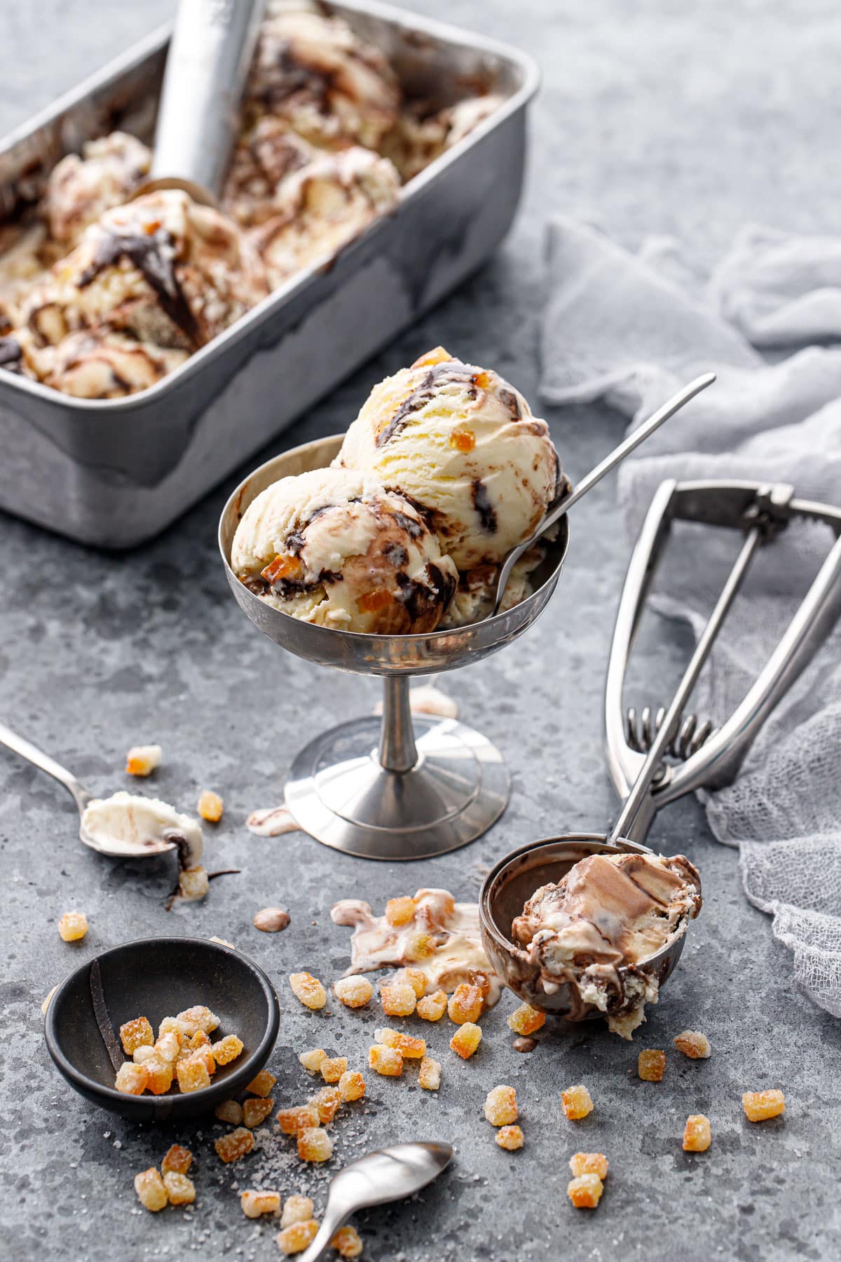 Messy scene with metal ice cream cup with two scoops of Candied Orange Ice Cream, ice cream scoop and bowl of candied orange pieces on the side, with a metal pan of scooped ice cream in the background.