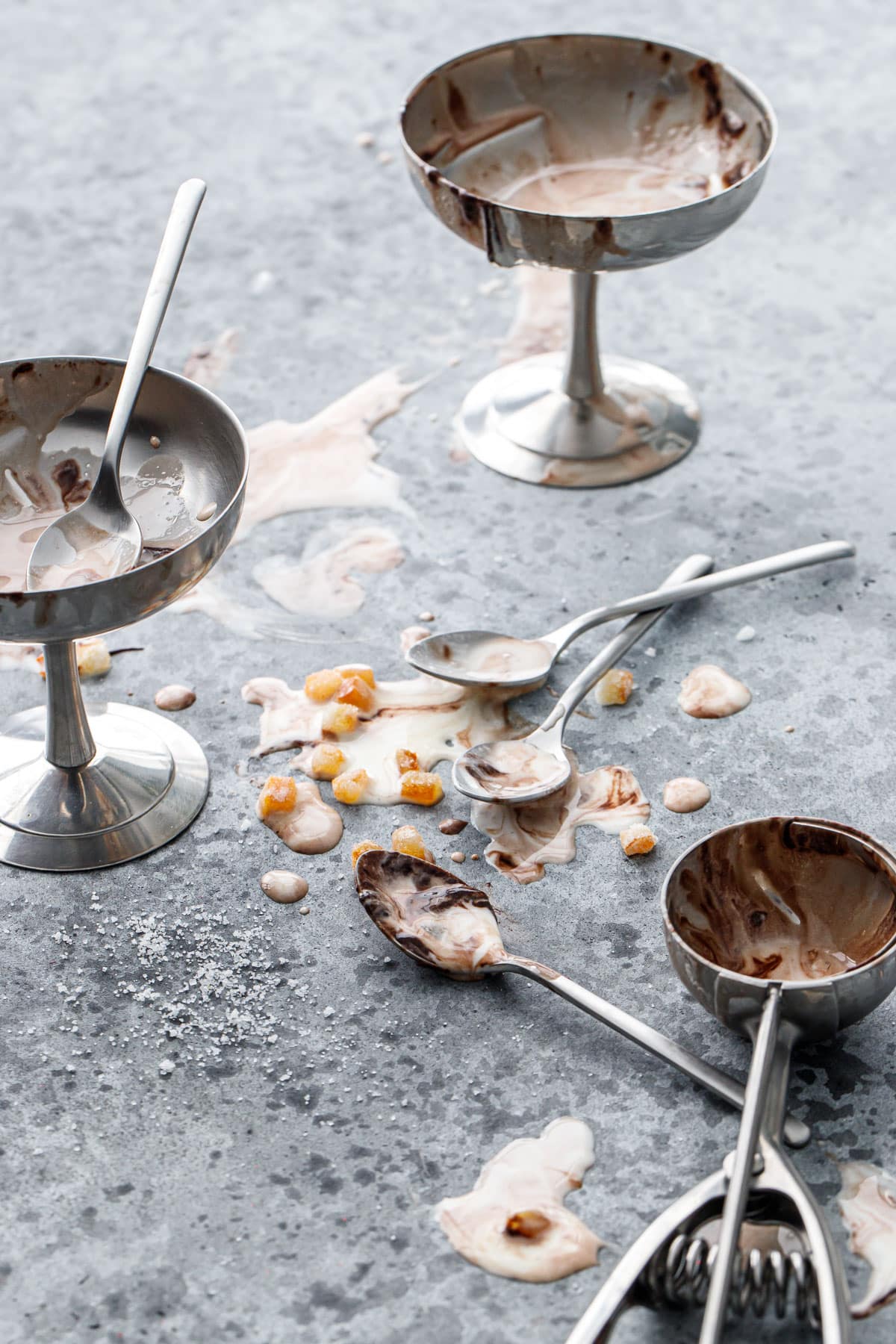 Messy scene with ice cream cups with melted ice cream, drippy spoons and bits of candied orange scattered about.