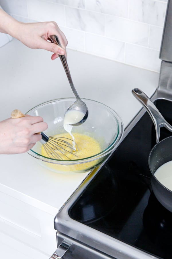 Ladling hot cream mixture into whisked egg yolks to temper it.