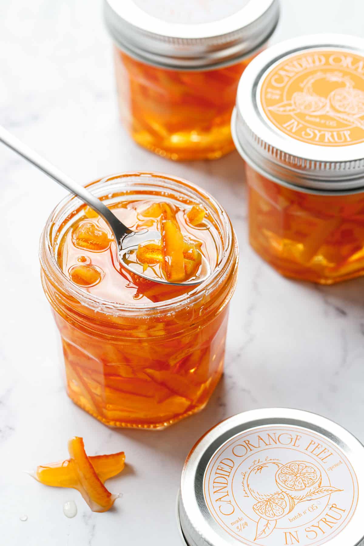 Backlit jars of Candied Orange Peel in Syrup, with a spoon and a few pieces of peel on the side.