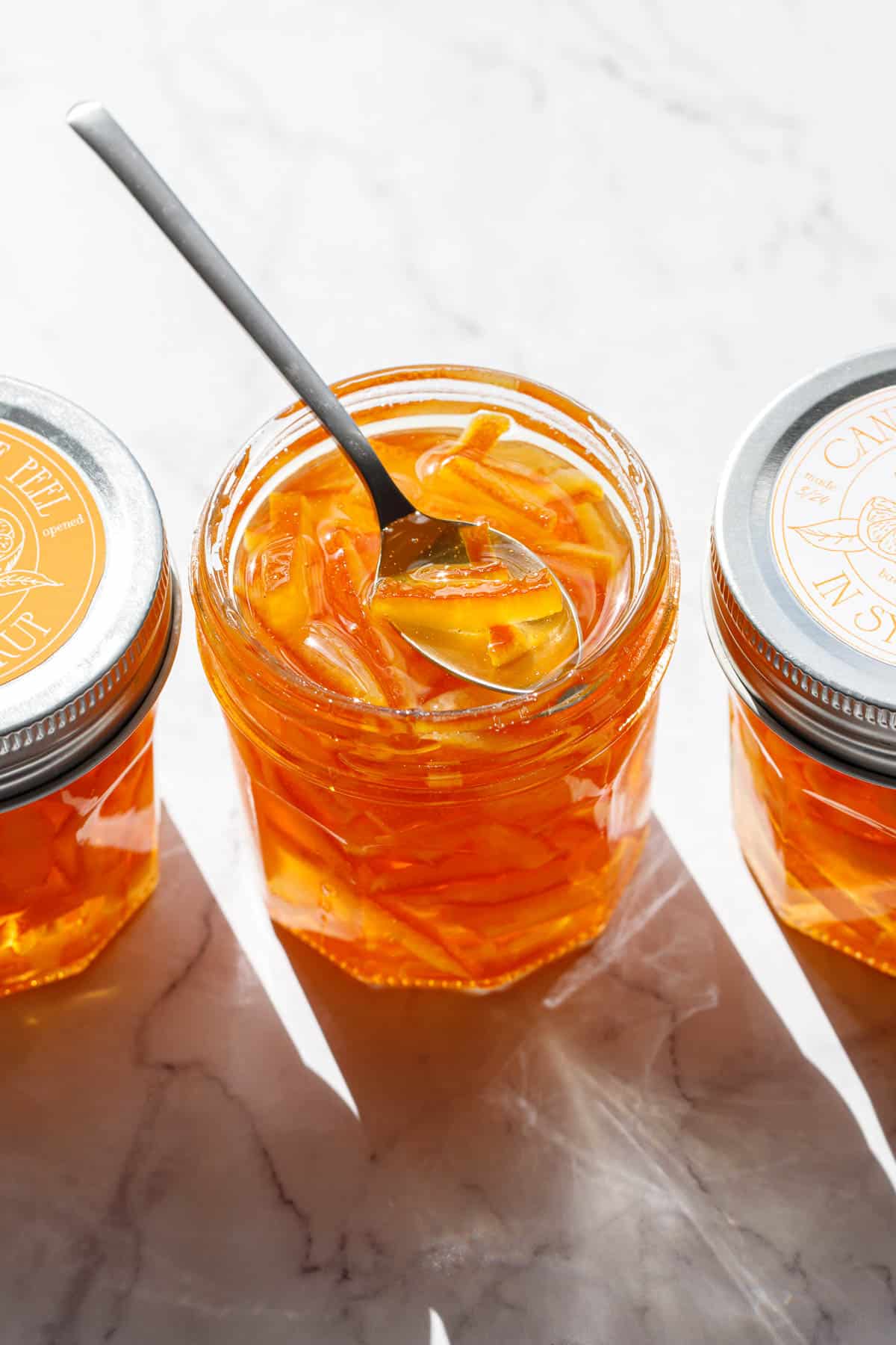 Three jars of Candied Orange Peel in Syrup, one jar open with a spoon resting on top, the other two jars sealed with designed orange and white labels on the lid.