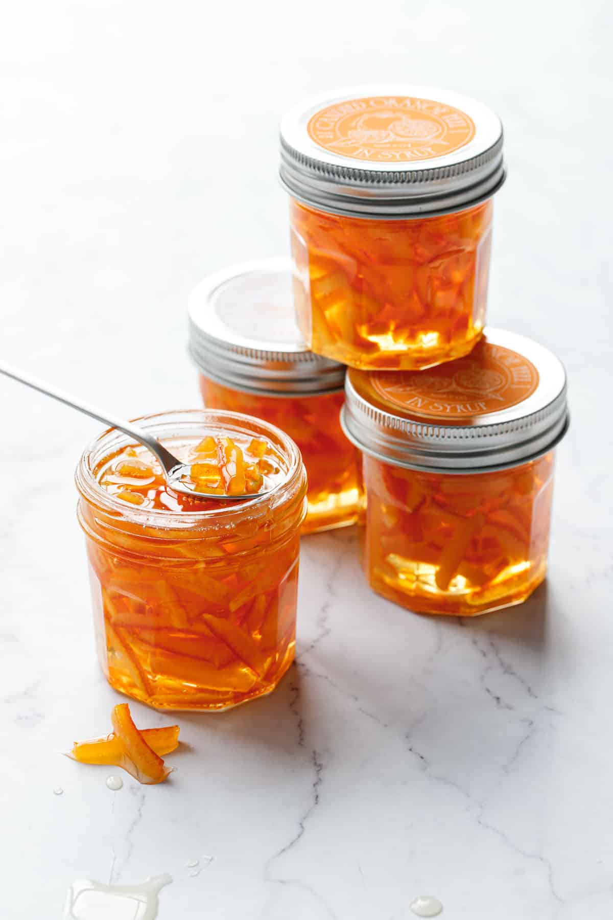 Stack of jars of Candied Orange Peel in Syrup, backlit and almost glowing from behind, with a few pieces of peel on a spoon and on the side.