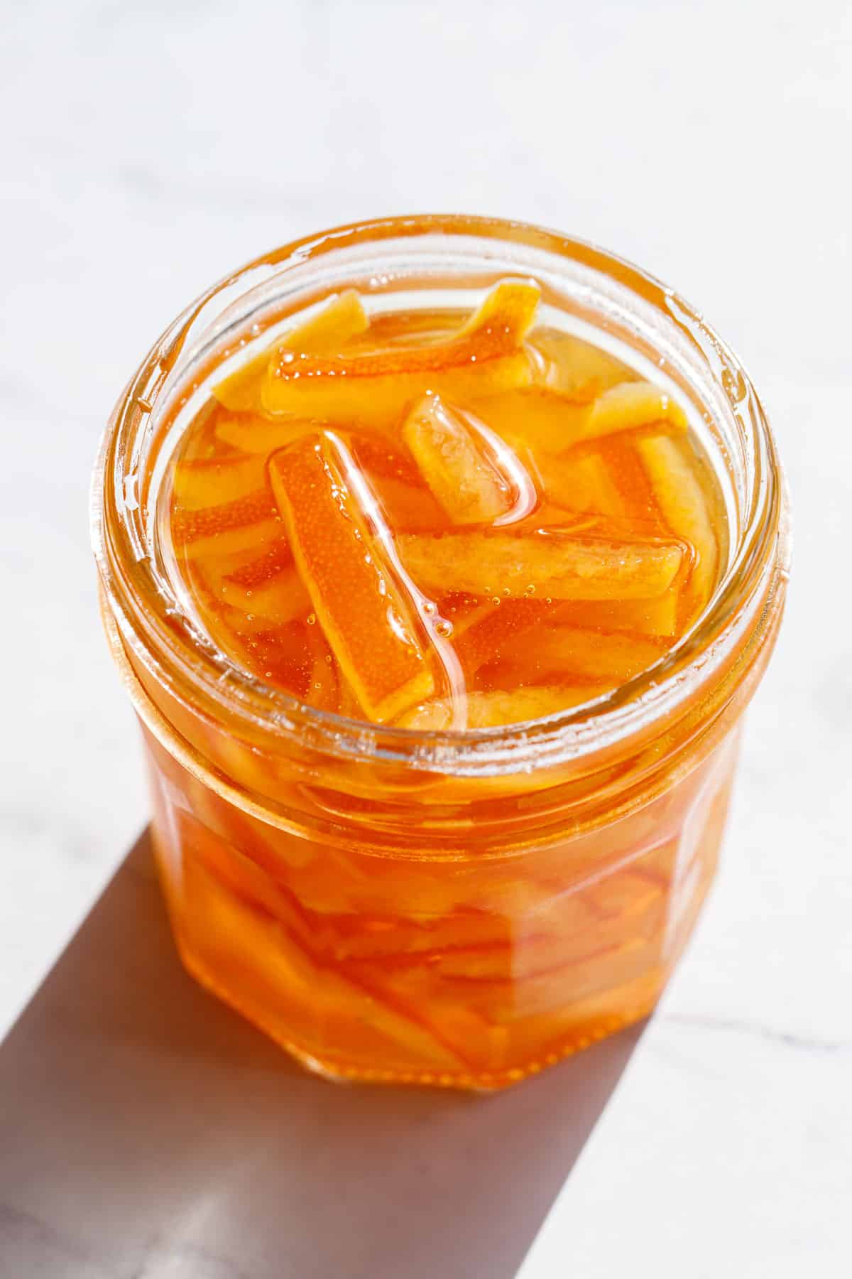 Direct light on an open glass canning jar of Candied Orange Peel in Syrup on a marble background.