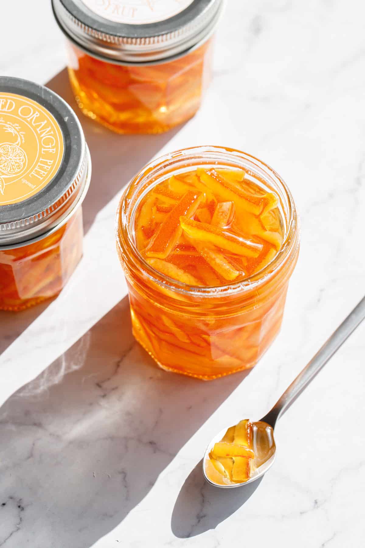 Three jars of Candied Orange Peel in Syrup in direct sunlight with harsh shadows and sparkling reflections off the shiny sugar syrup, spoon with a few pieces of candied peel on the side.