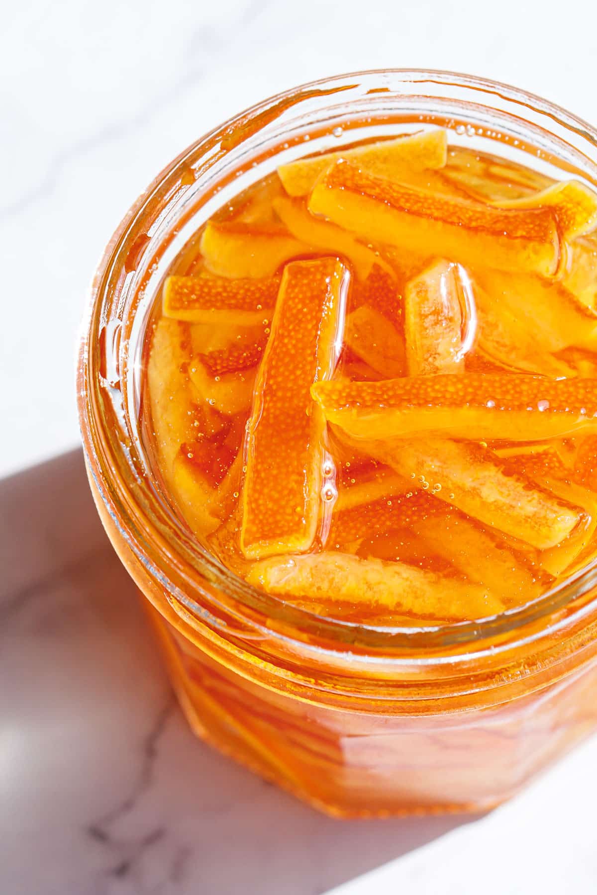 Closeup, open glass canning jar filled  with Candied Orange Peel in Syrup, showing the texture of the candied orange peel and crystal clear sugar syrup in direct sunlight.