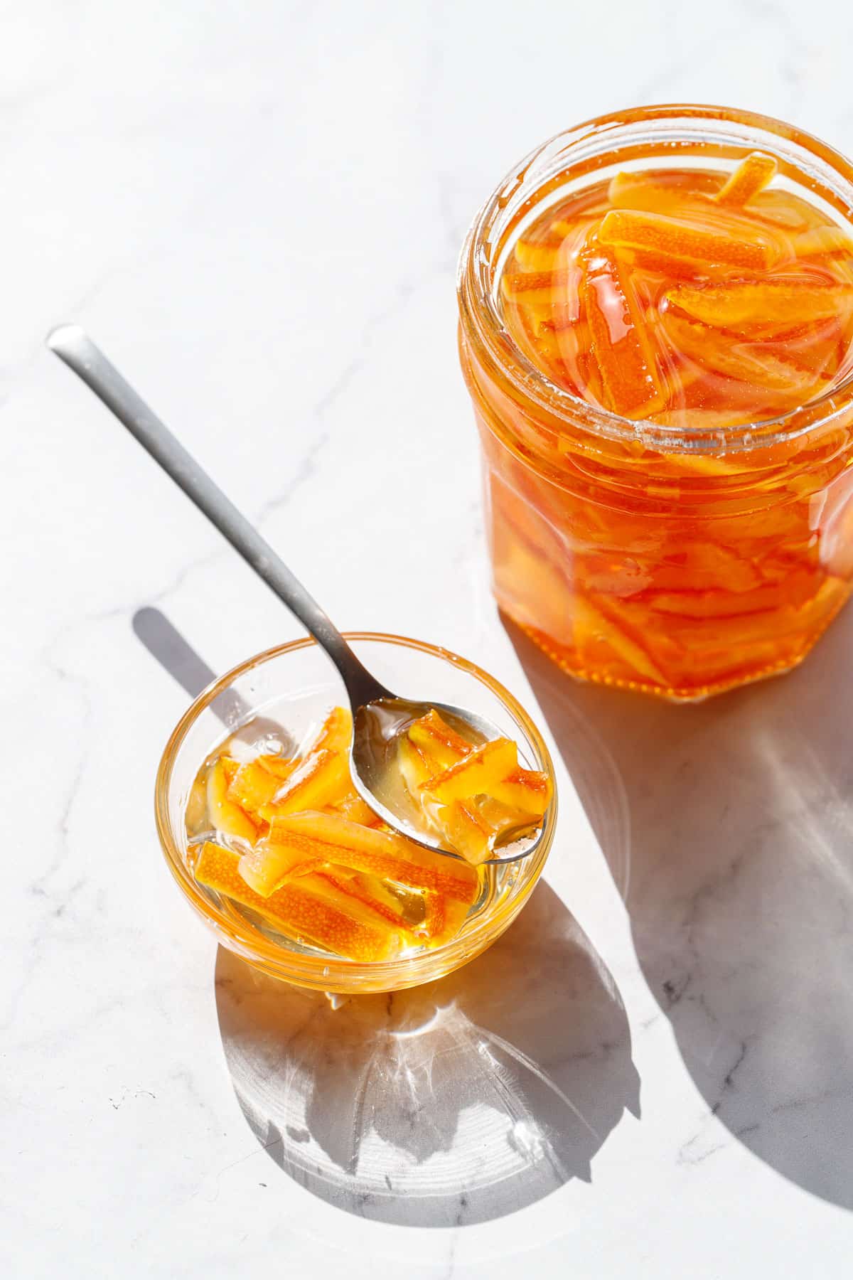 Bowl and open glass jar filled with Candied Orange Peel in Syrup, with a spoon with a few pieces of candied peel balanced on top of the bowl.