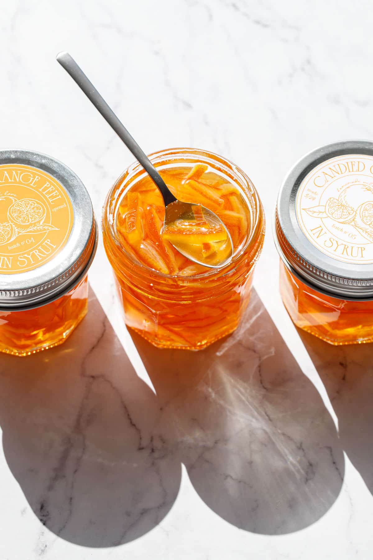 Three jars of Candied Orange Peel in Syrup, one jar open with a spoon resting on top, the other two jars sealed with designed orange and white labels on the lid.