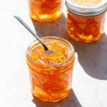 Faceted glass canning jars filled with Candied Orange Peel in Syrup in direct sunlight with harsh shadows and highlights on a marble background.