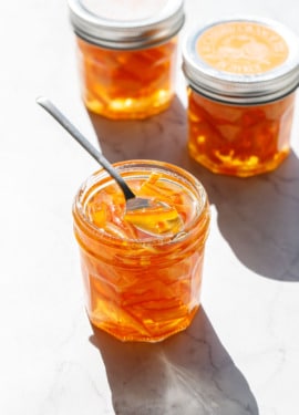 Faceted glass canning jars filled with Candied Orange Peel in Syrup in direct sunlight with harsh shadows and highlights on a marble background.