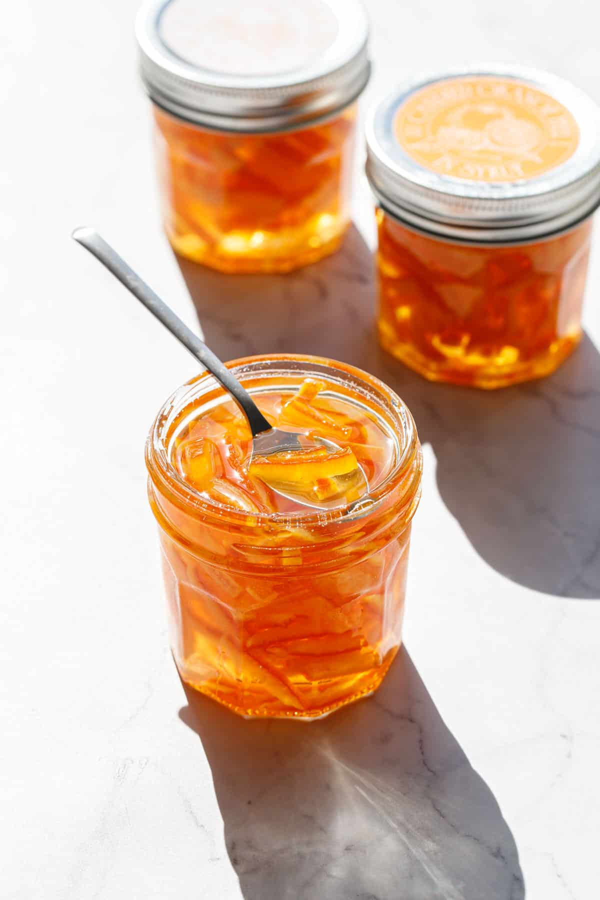 Faceted glass canning jars filled with Candied Orange Peel in Syrup in direct sunlight with harsh shadows and highlights on a marble background.