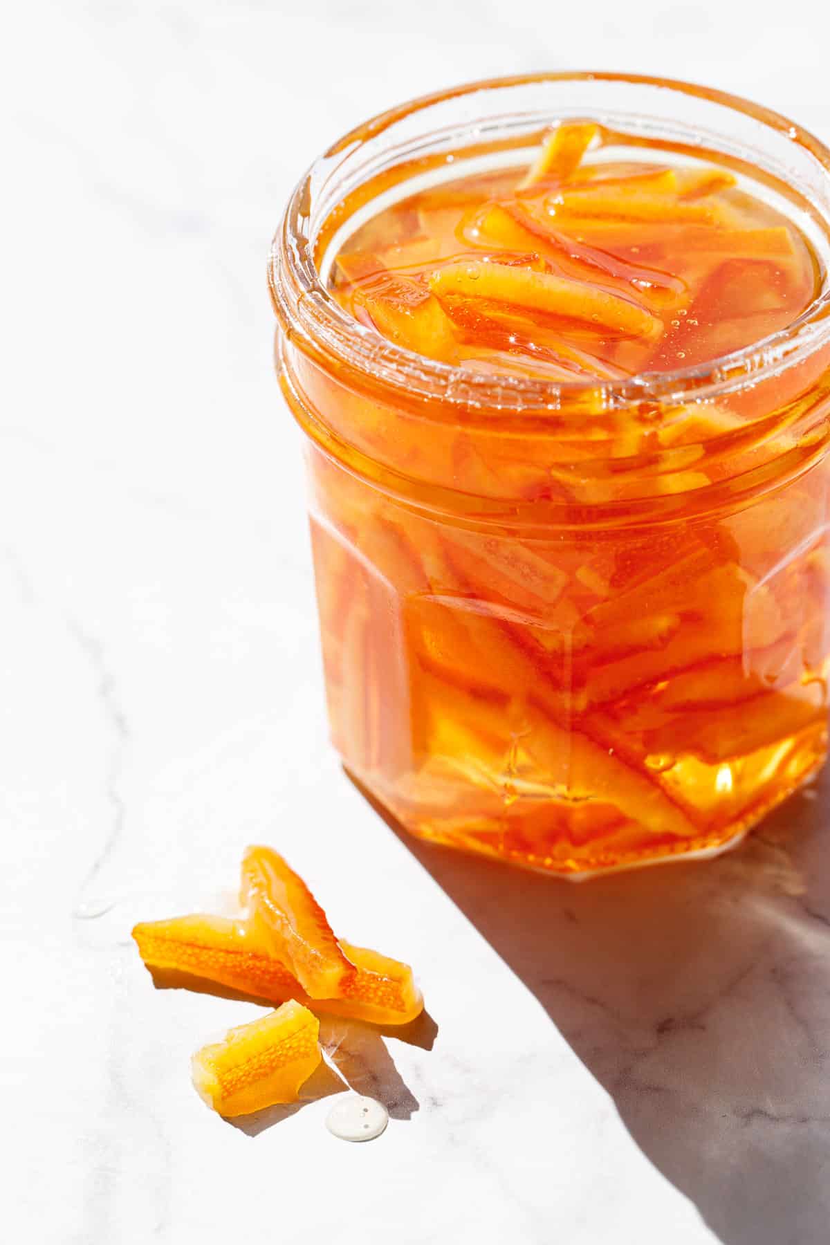 Closeup, open glass jar of Candied Orange Peel in Syrup with a few pieces of peel sitting on the marble background next to the jar.