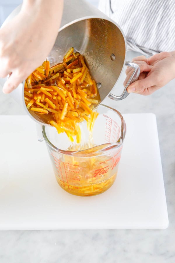 Pouring candied orange peel from saucepan into a spouted glass measuring cup.