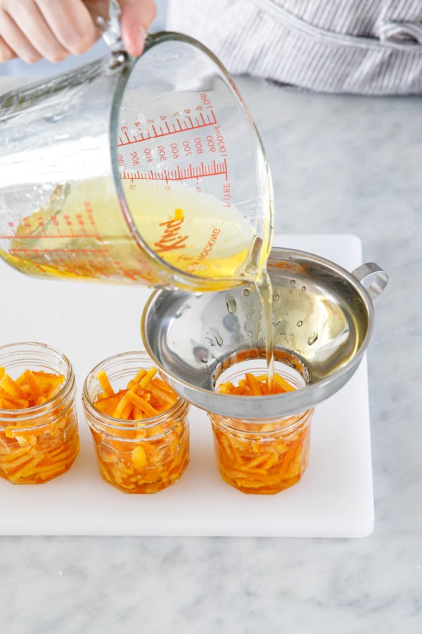 Pouring candied orange syrup on top of orange peel pieces in jars until covered in syrup.