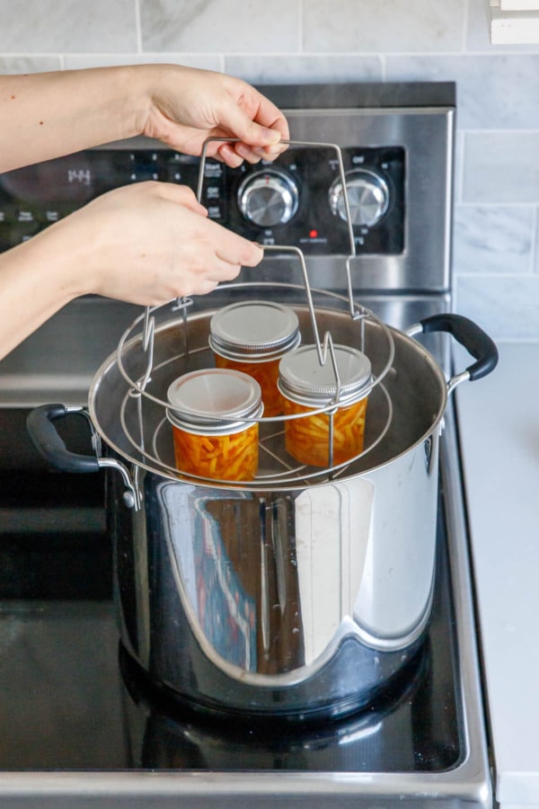 Lowering canning rack with jars of candied orange peel into a large stainless pot.