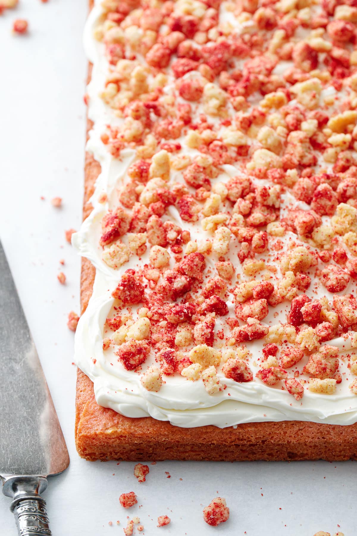 Strawberry Crunch Sheet Cake, freshly frosted with buttermilk frosting and sitting on a piece of parchment with a cake server and messy crumbs scattered around it.