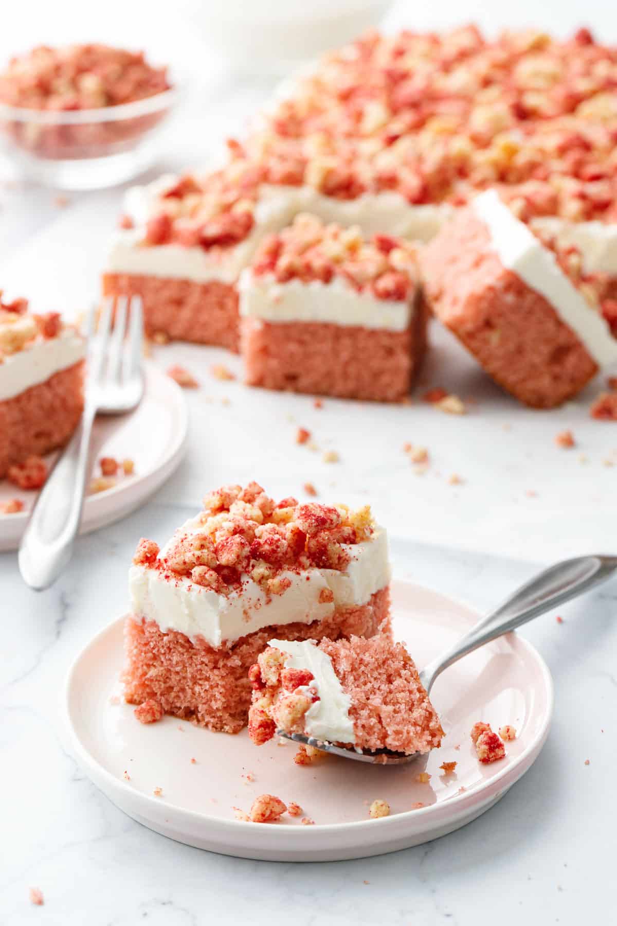 Slice of Strawberry Crunch Sheet Cake with Buttermilk Frosting on a dessert plate, with a forkful of cake on the side showing the texture, full cake out of focus in the background.