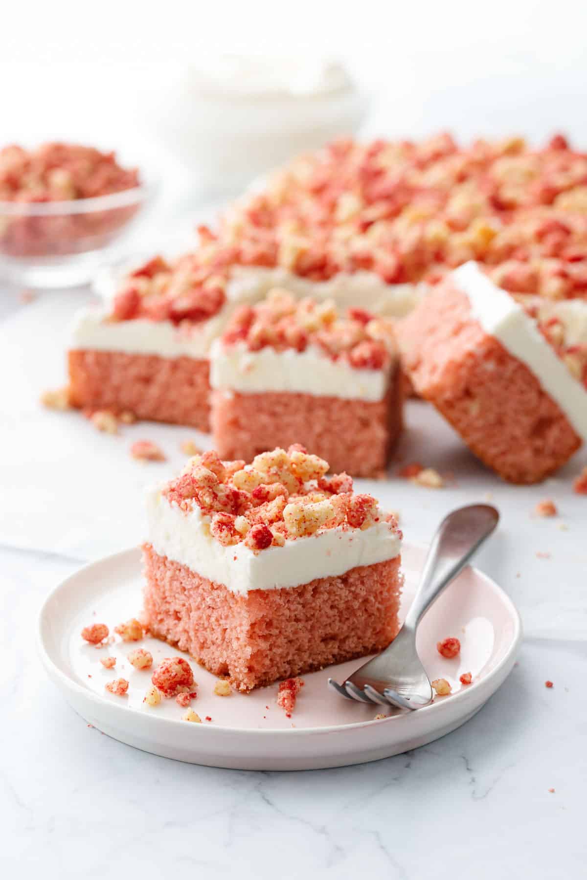 Strawberry Crunch Sheet Cake with Buttermilk Frosting, focus on one piece of cake sitting on a pink dessert plate with a fork alongside and messy crumbs, and a bowl of more crumbs and frosting out of focus in the background.