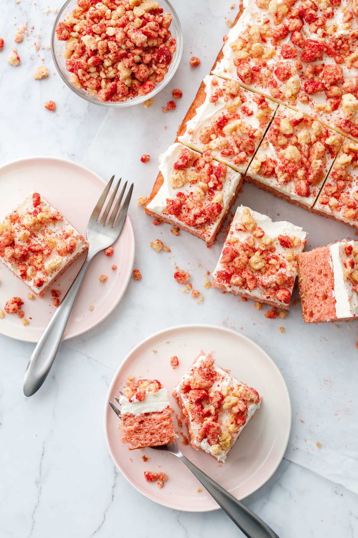 Overhead, messy scene with cut pieces of Strawberry Crunch Sheet Cake with Buttermilk Frosting on pink dessert plates, marble background, and a bowl of more strawberry crunch crumbs on the side.