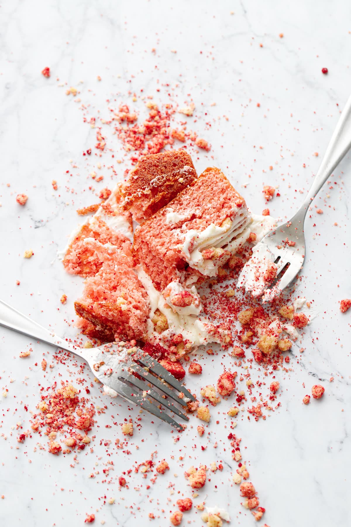 Messy shot of smashed strawberry crunch cake on a marble background, two forks smeared with cake and frosting and scattered crumbs.