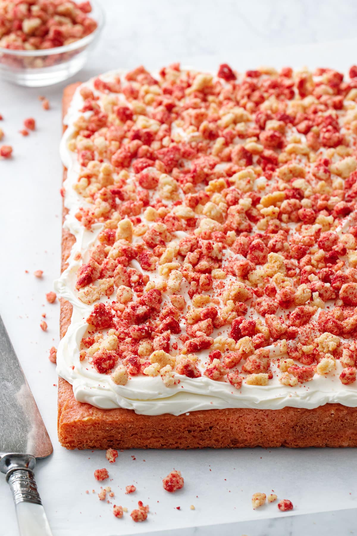 Strawberry Crunch Sheet Cake, freshly frosted with buttermilk frosting and sitting on a piece of parchment with a cake server on the side, bowl of strawberry crunch topping in the background, and messy crumbs scattered around it.