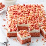 Strawberry Crunch Sheet Cake with Buttermilk Frosting cut into serving squares, on a piece of parchment with messy crumbs, cake server, and a bowl of more crumbs and frosting in the background.