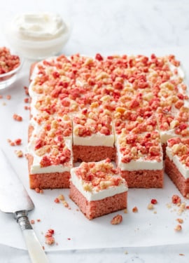 Strawberry Crunch Sheet Cake with Buttermilk Frosting cut into serving squares, on a piece of parchment with messy crumbs, cake server, and a bowl of more crumbs and frosting in the background.