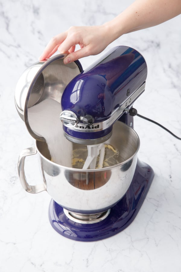 Pouring sugar into the bowl of a blue stand mixer with creamed butter.