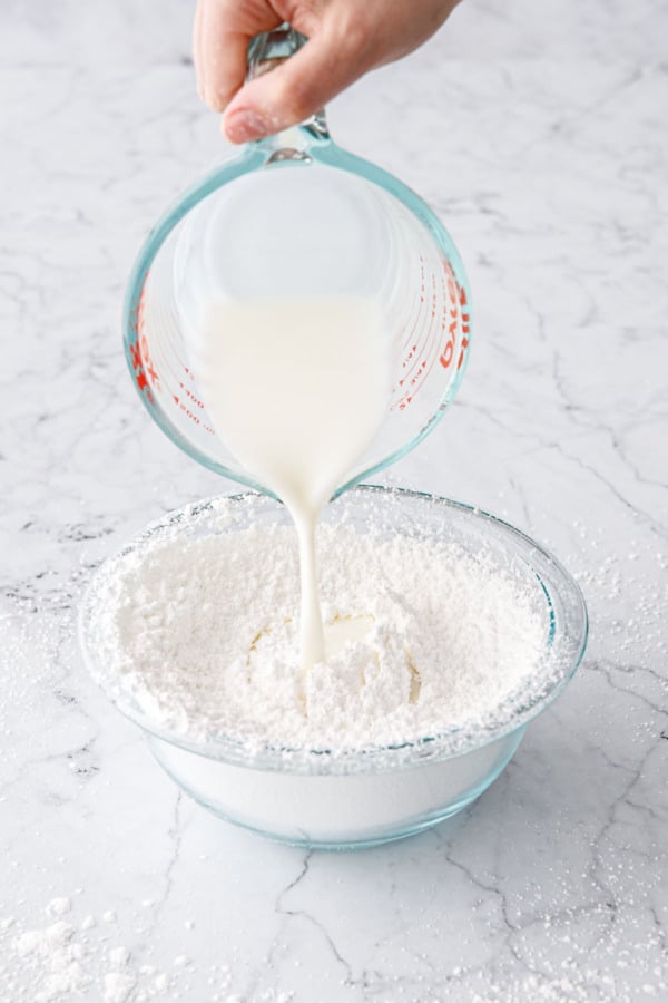 Pouring buttermilk and heavy cream into glass mixing bowl with sifted powdered sugar.