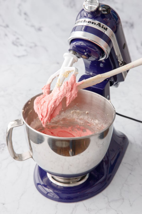 Spatula scraping the beater and sides of mixing bowl filled with pink strawberry cake batter.