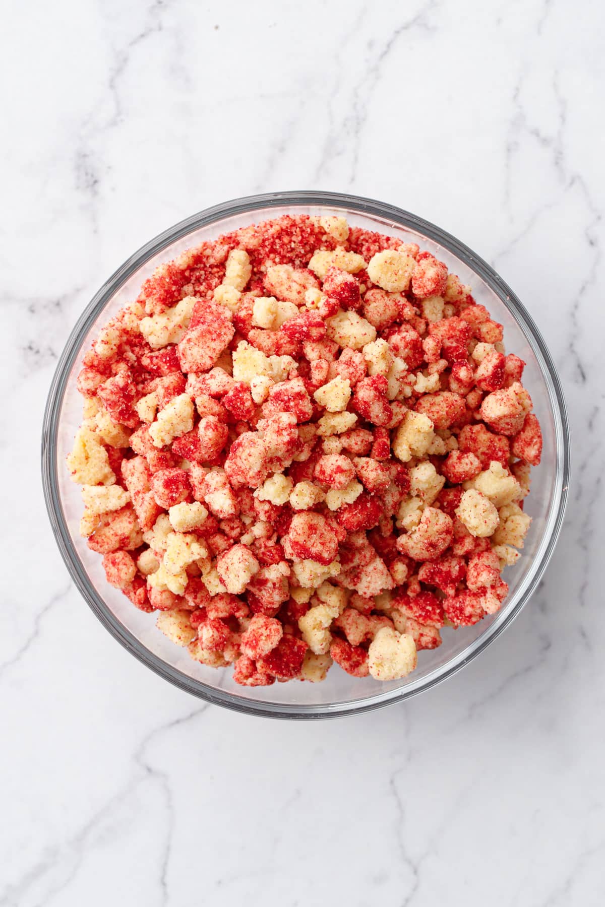 Overhead, glass bowl filled with red and yellow bits of Strawberry Shortcake Crunch Topping on a marble background.