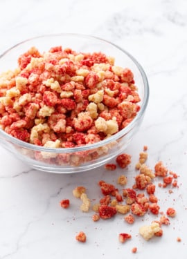 Glass bowl of Strawberry Shortcake Crunch Topping on a marble background, with a few crumbs scattered alongside the bowl.