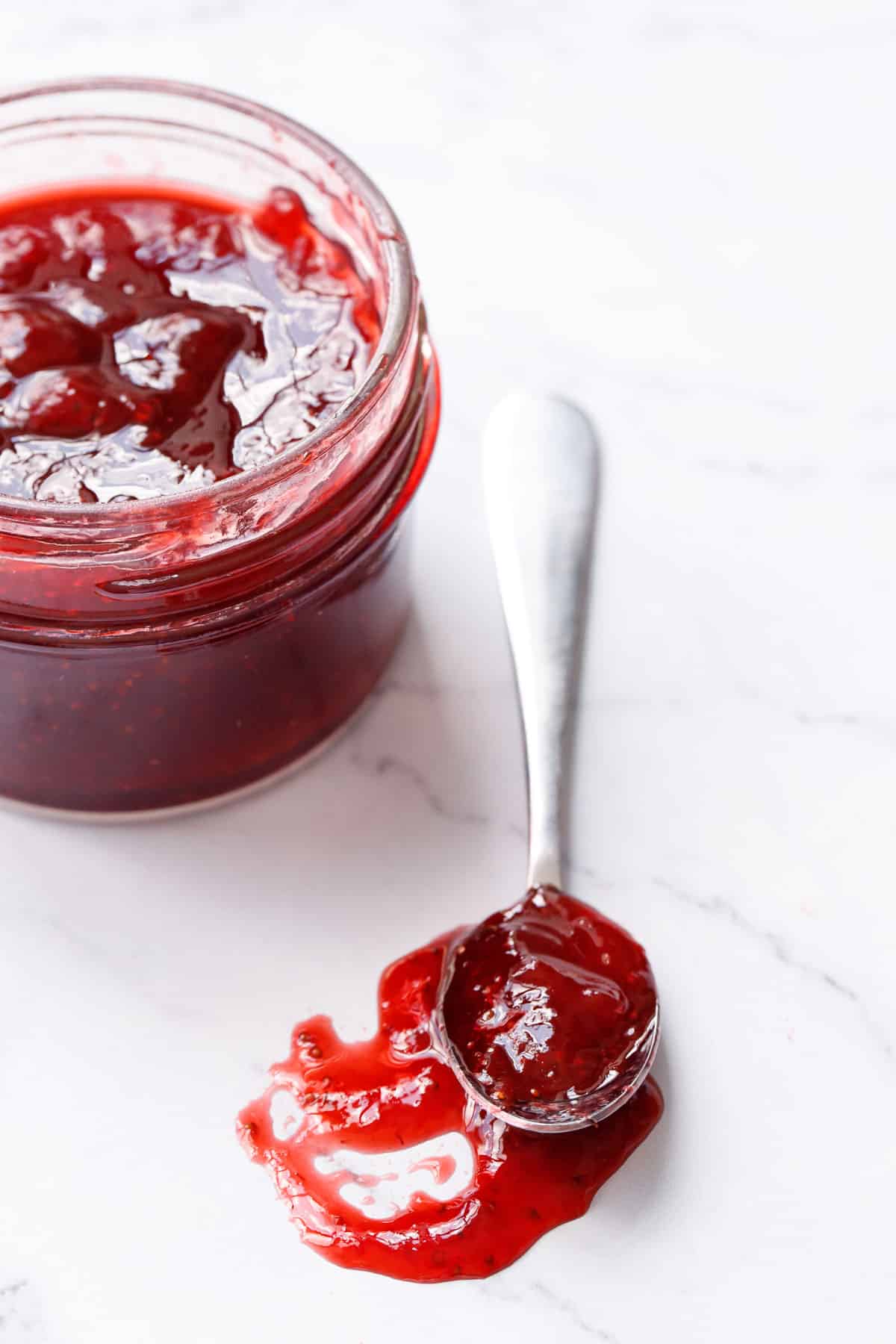 Jar and spoonful of Old Fashioned strawberry jam on a marble background, close up to show the jammy texture and rich color.