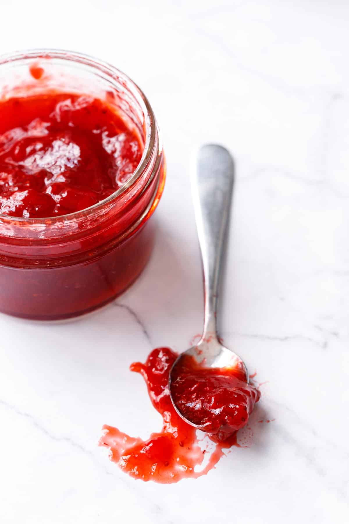 Jar and spoonful of classic strawberry jam on a marble background, close up to show the jammy texture and bright red color.
