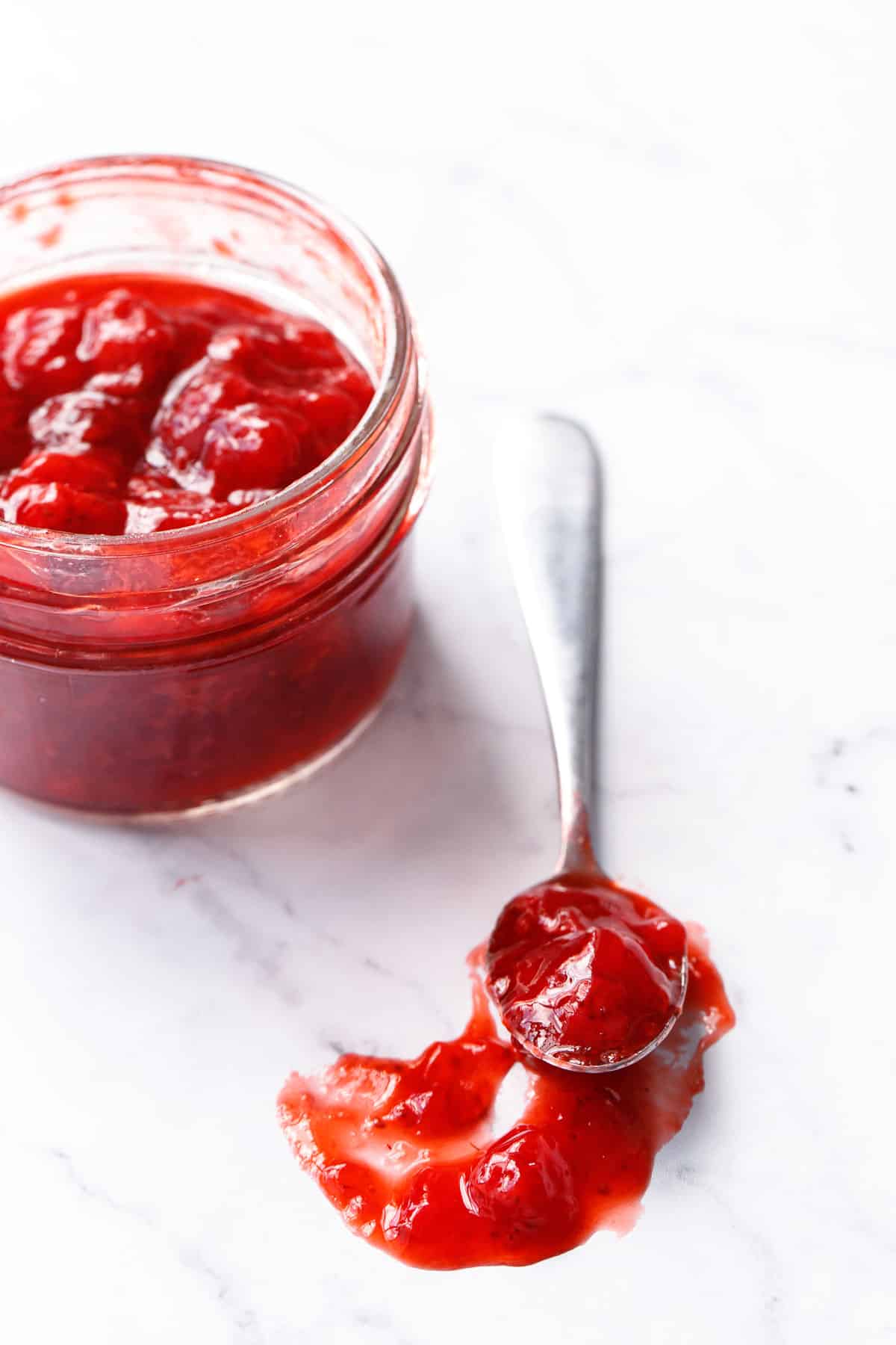 Jar and spoonful of low-sugar strawberry jam on a marble background, close up to show the fruit-forward texture and bright color.