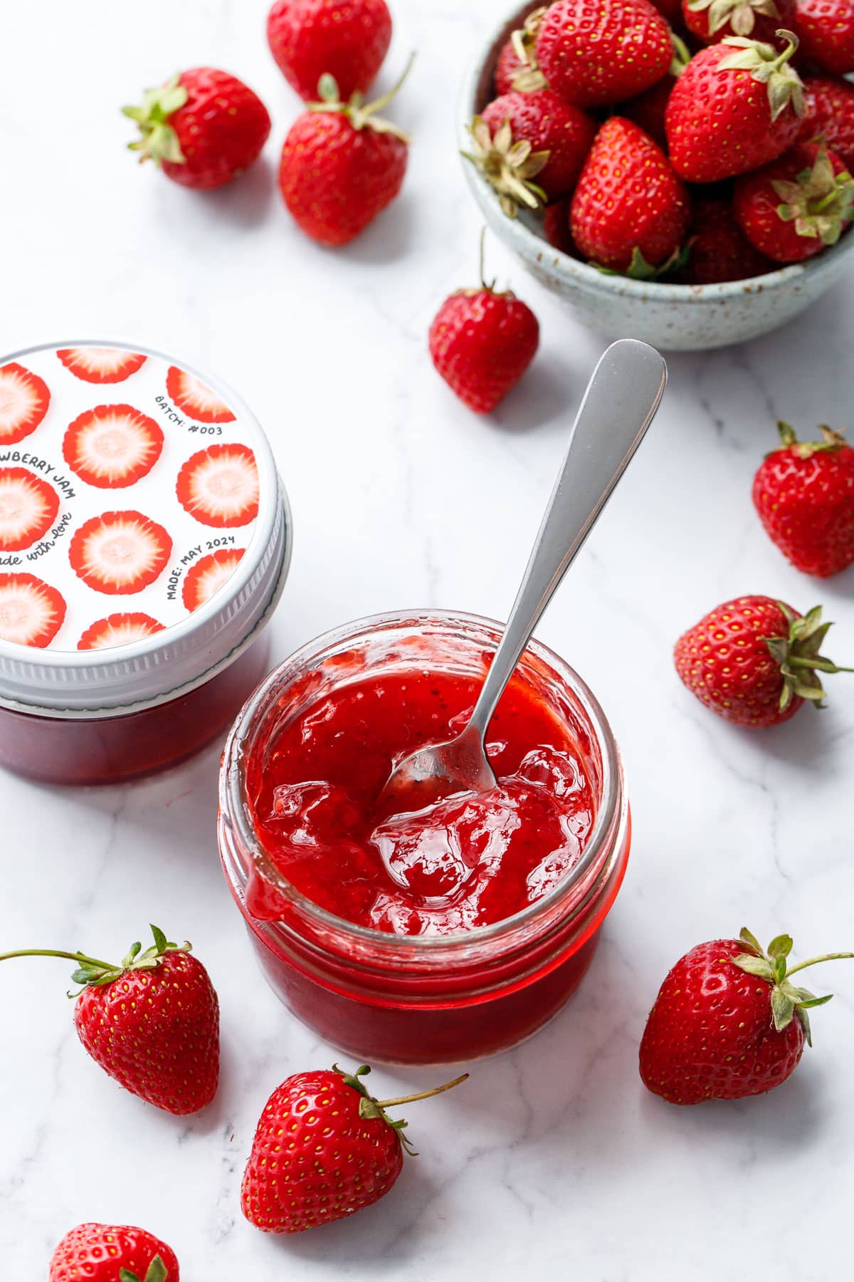 Classic Strawberry Jam in a glass jar with fresh strawberries on a marble background, one jar open in the foreground with a spoon in it to show the texture.