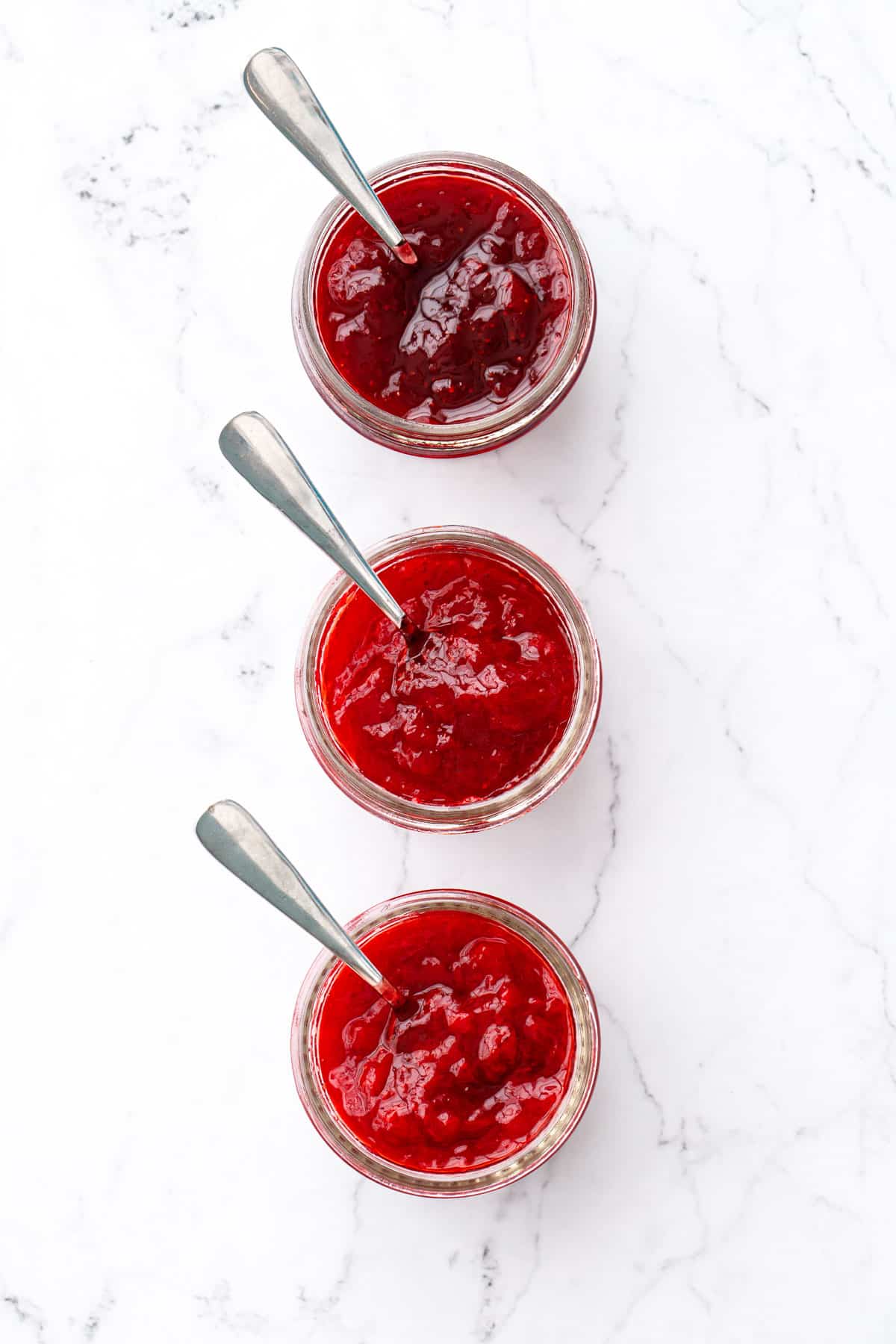 Overhead, three open glass jars with spoons on a marble background, comparing the color/texture of 3 different kinds of strawberry jam.