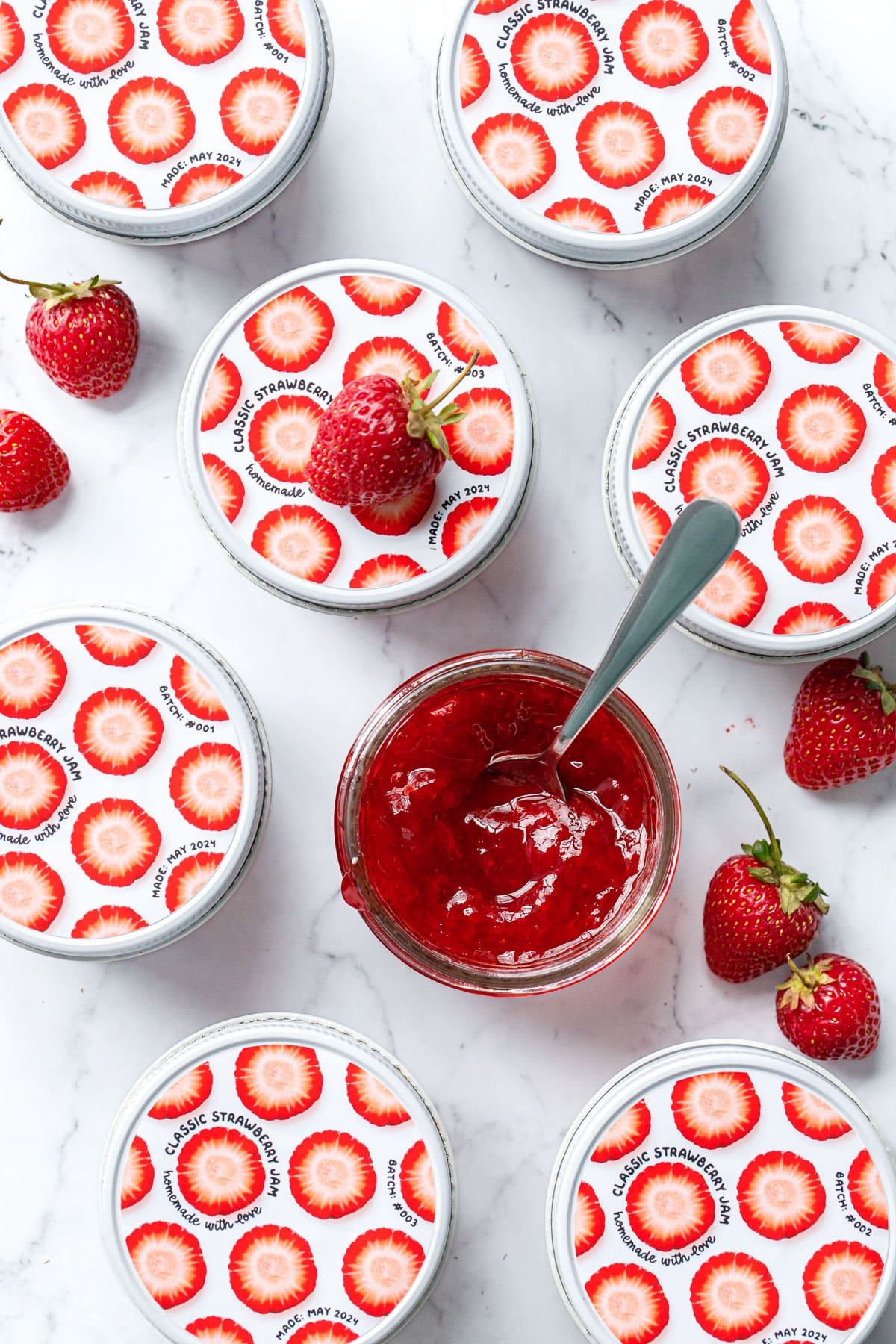 Overhead, jars of Classic Strawberry Jam on a marble background, one jar open with a spoon in it, the others closed with cute designed labels visible on the lids.