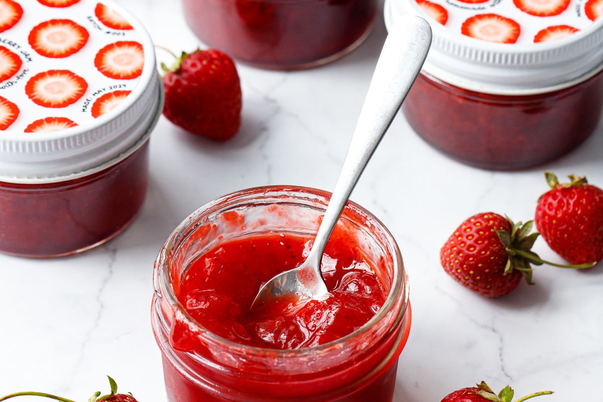 Classic Strawberry Jam in a glass jar with fresh strawberries on a marble background, one jar open in the foreground with a spoon in it to show the texture.