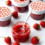 Classic Strawberry Jam in a glass jar with fresh strawberries on a marble background, one jar open in the foreground with a spoon in it to show the texture.