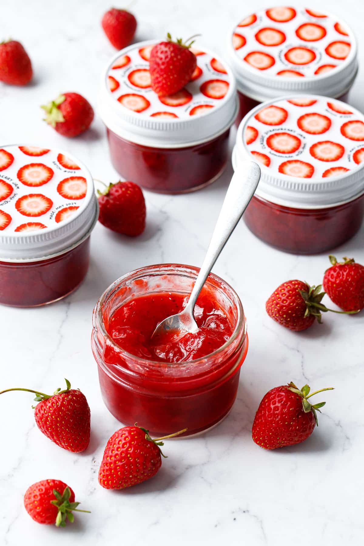 Classic Strawberry Jam in a glass jar with fresh strawberries on a marble background, one jar open in the foreground with a spoon in it to show the texture.