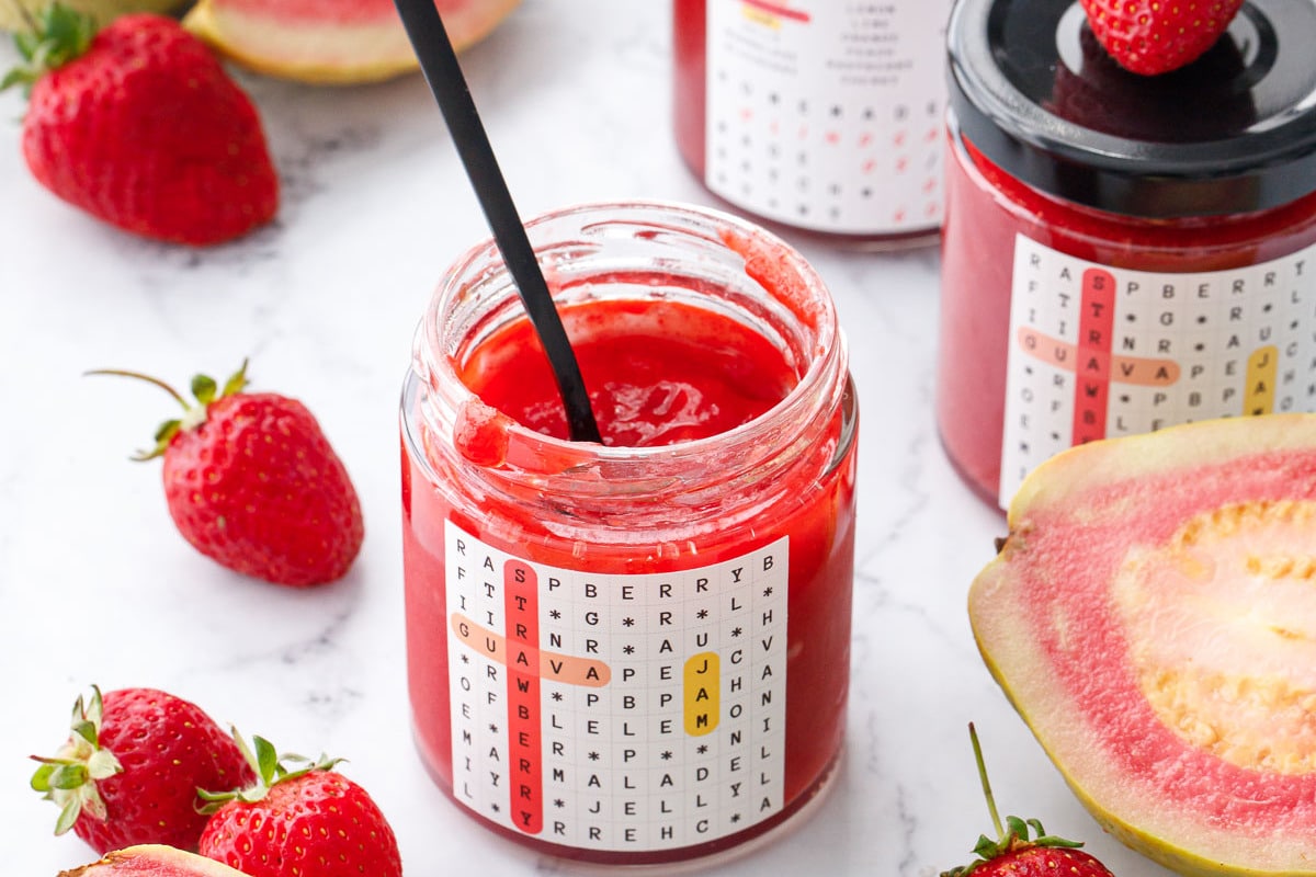 Jars of Guava Strawberry Jam on a marble background with fresh strawberries and whole and cut pink guavas scattered around.