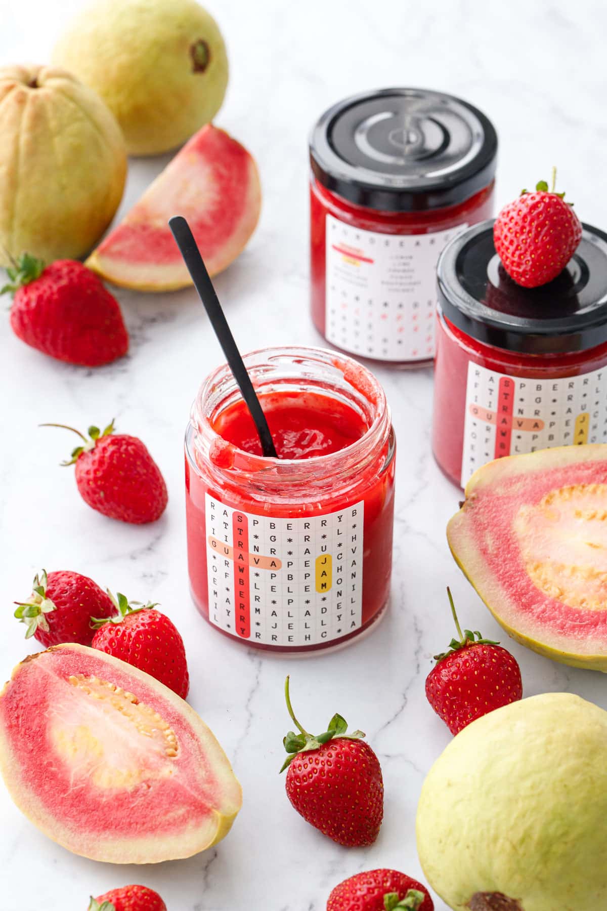 Jars of Guava Strawberry Jam on a marble background with fresh strawberries and whole and cut pink guavas scattered around.