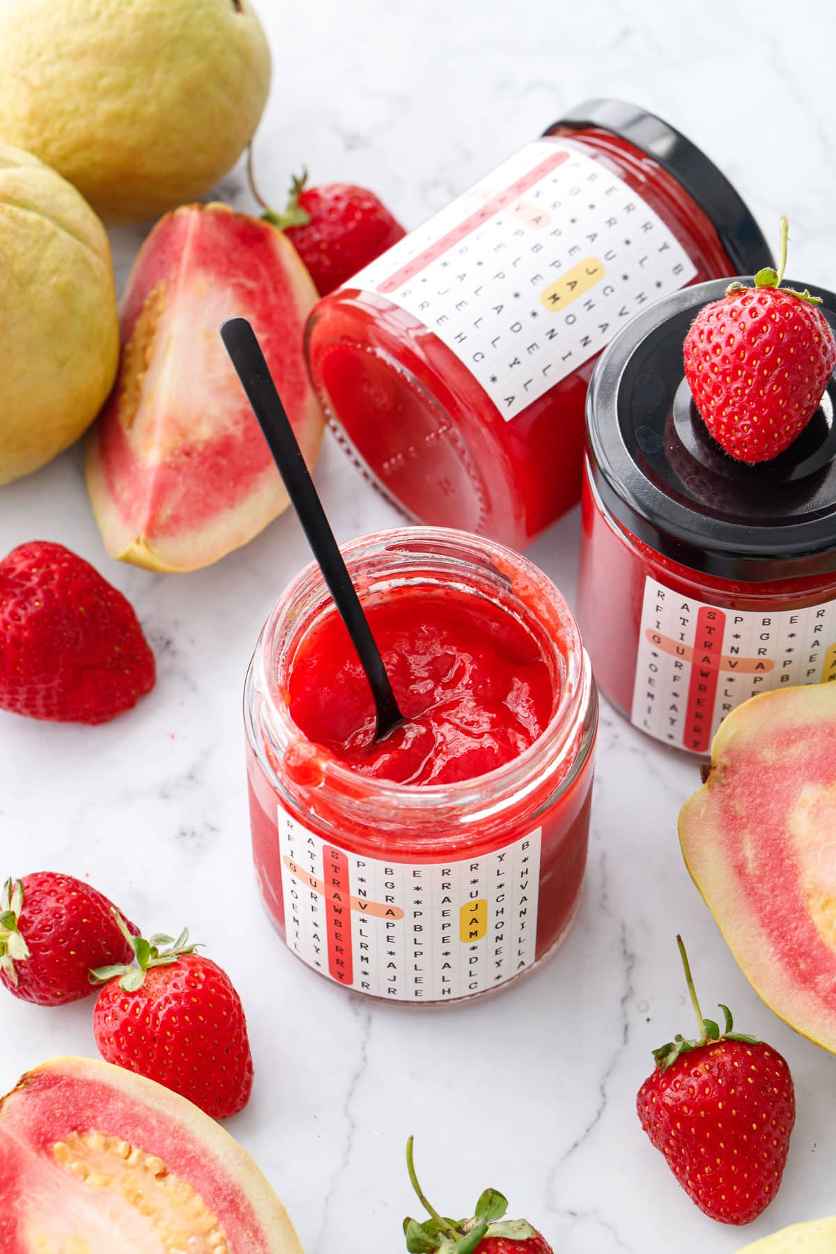 Jars of Guava Strawberry Jam on a marble background with fresh strawberries and whole and cut pink guavas scattered around.