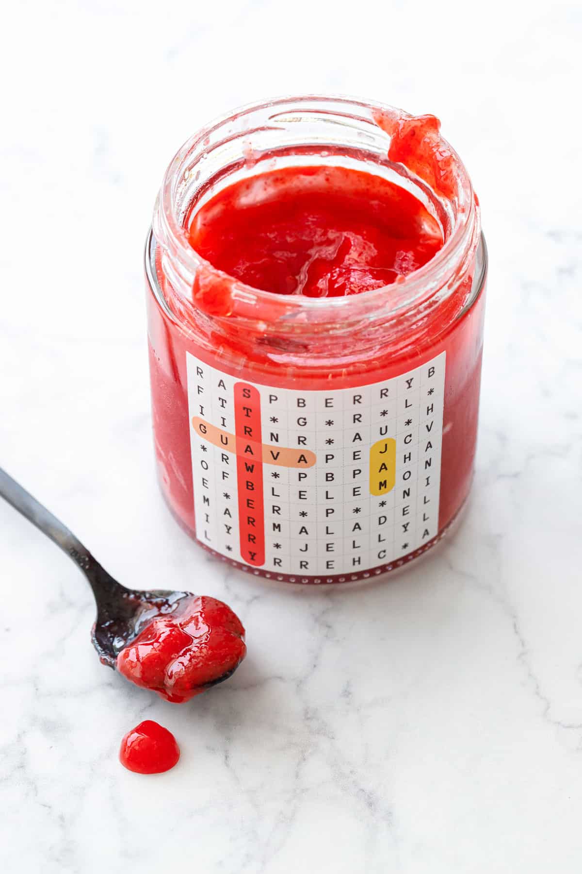 Jar of Guava Strawberry Jam on a marble background with a black spoon with dollop of jam dripping on the side.