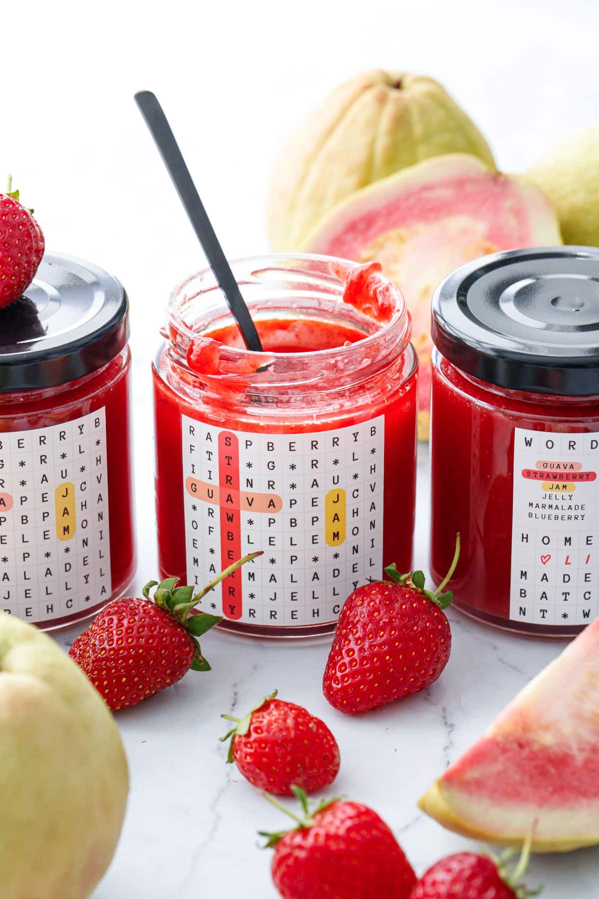 Jars of Guava Strawberry Jam, one opened with as spoon in the jar, with fresh strawberries and pink guavas in the background.