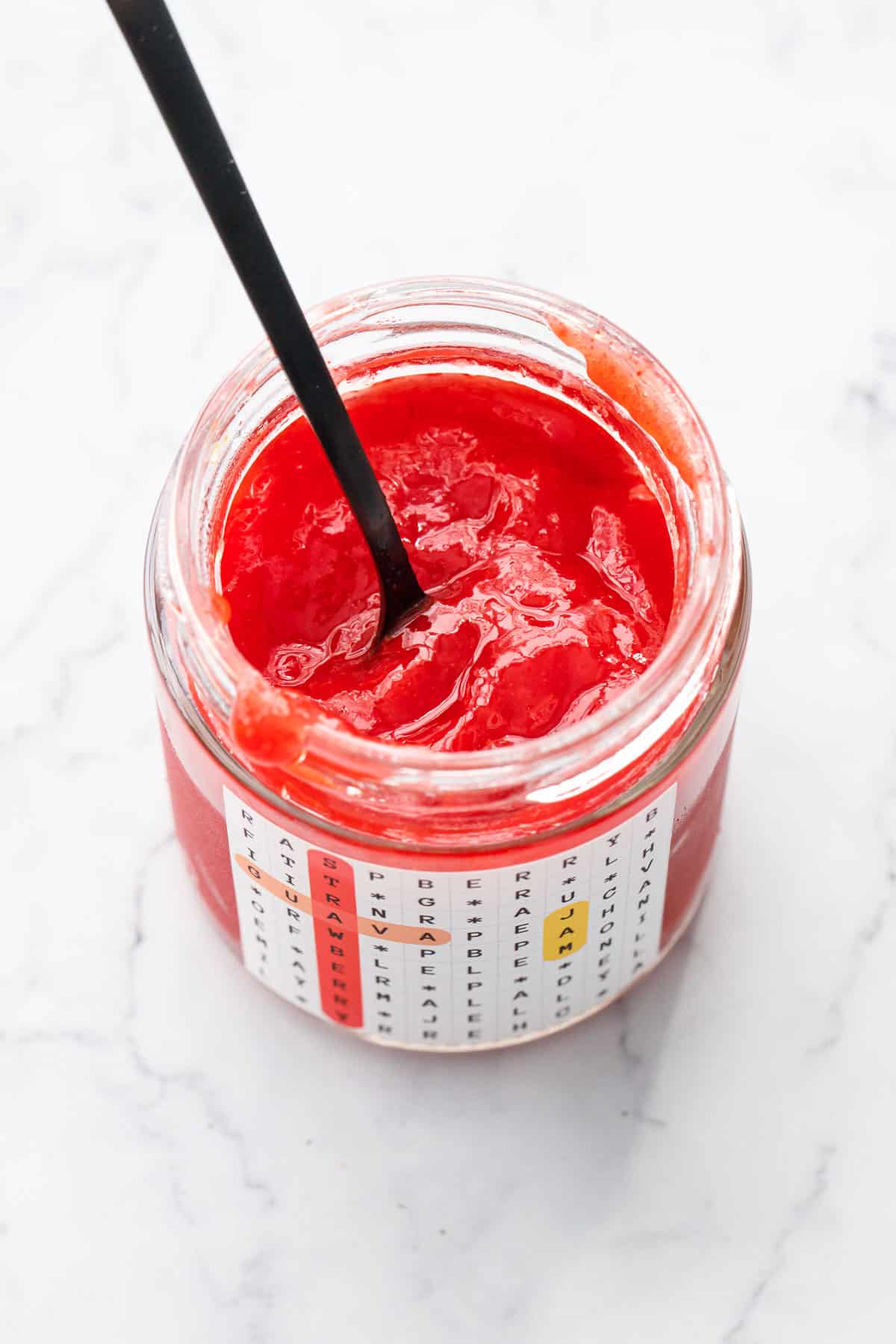 Looking in to an open jar of Strawberry & Guava Jam with a black spoon, showing the bold color, smooth texture.