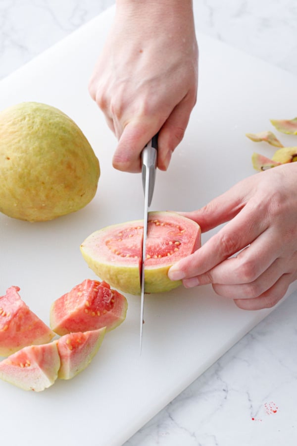 Cutting pink guavas in half on a cutting board with a knife.