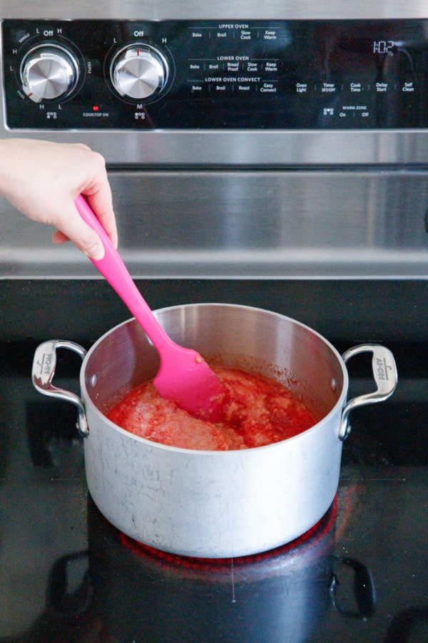 Stirring fruit in a saucepan on the stovetop with a pink spatula.