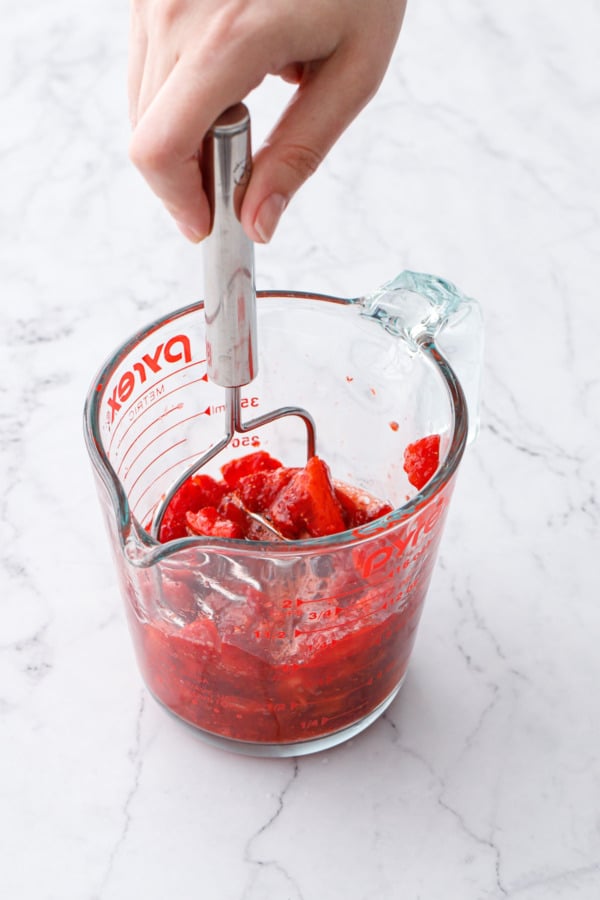 Mashing chopped ripe strawberries in a glass measuring cup with a small potato masher.