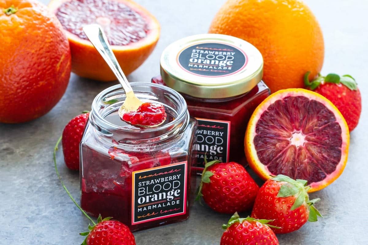 Jars of strawberry blood orange marmalade on a gray background with fresh oranges and strawberries.